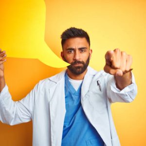 Young indian doctor man holding speech bubble standing over isolated yellow background pointing with finger to the camera and to you, hand sign, positive and confident gesture from the front